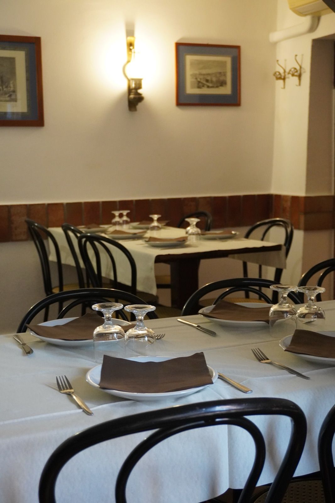 La sala apparecchiata: tovaglie bianche, sedie di legno curvato, le stampe alle pareti e le piastrelle marroni